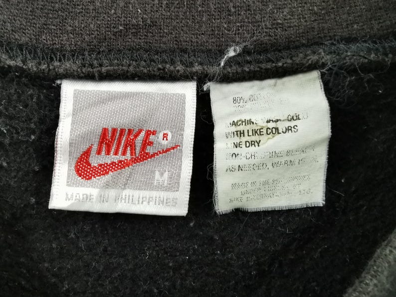 K&ouml;nnte beinhalten: Nahaufnahme eines schwarzen Kleidungsst&uuml;cks mit zwei Etiketten. Das linke Etikett zeigt das rote Nike-Logo und das Wort "NIKE" in Wei&szlig;. Das rechte Etikett enth&auml;lt Waschanweisungen. Das Kleidungsst&uuml;ck wird in den Philippinen hergestellt.