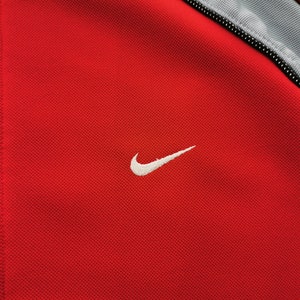 Op de afbeelding: Close-up van een rode sportjas met een wit Nike-logo op de borst. De jas heeft een volledige ritssluiting en grijze accenten op de schouders en kraag. De stof heeft een getextureerde uitstraling.