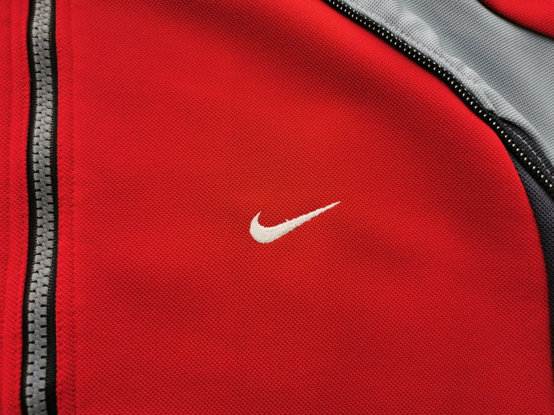 Op de afbeelding: Close-up van een rode sportjas met een wit Nike-logo op de borst. De jas heeft een volledige ritssluiting en grijze accenten op de schouders en kraag. De stof heeft een getextureerde uitstraling.