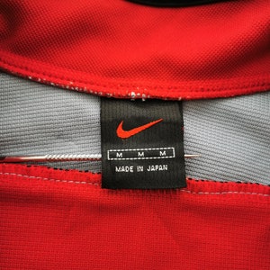 Op de afbeelding: Close-up van een rode Nike polokraag met een grijze binnenkant. Een zwart label met het Nike swoosh-logo en "MADE IN JAPAN" is zichtbaar. Het label heeft ook drie "M" maatlabels.