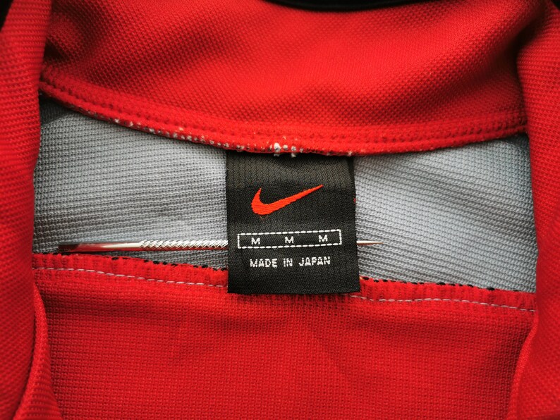 Op de afbeelding: Close-up van een rode Nike polokraag met een grijze binnenkant. Een zwart label met het Nike swoosh-logo en "MADE IN JAPAN" is zichtbaar. Het label heeft ook drie "M" maatlabels.