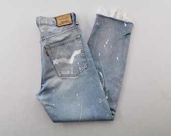 Puede incluir: Vaqueros de mezclilla azul claro con salpicaduras de pintura blanca, dobladillo deshilachado y una etiqueta de cuero marrón de Levi Strauss & Co. en la cintura. El bolsillo trasero muestra un diseño arqueado pintado en blanco.