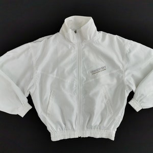 Puede incluir: Chaqueta blanca con cremallera completa, cuello alzado y puños y dobladillo elásticos. La chaqueta presenta el texto "CHRISEVERT INTERNATIONAL CLOTHING" en gris en el pecho izquierdo. La chaqueta es de material ligero.