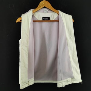 Puede incluir: Chaleco color crema con forro de malla morada clara, colgado de una percha de madera. El chaleco est&aacute; abierto, revelando el forro interior. La marca "DIAPLEX" es visible en la etiqueta. El fondo es de color oscuro.
