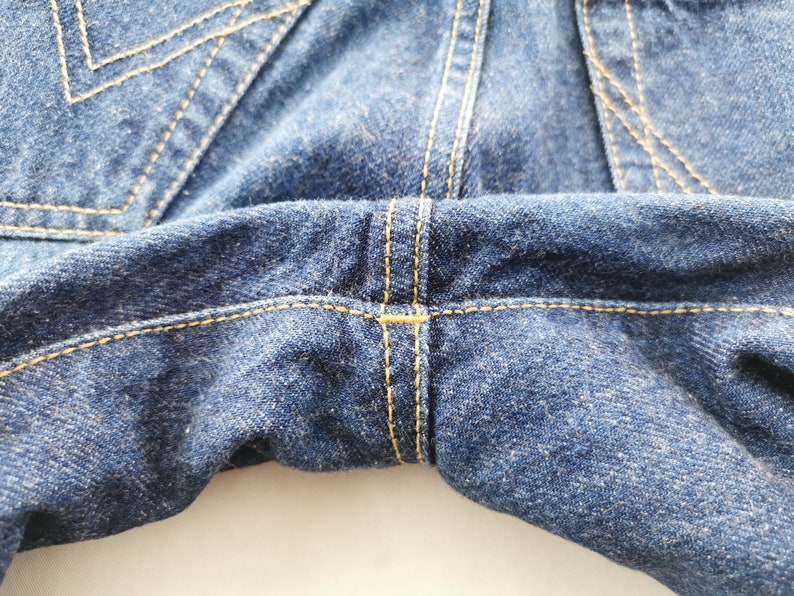May include: Close-up of blue denim jeans, showcasing the stitching details. The image highlights the seams and the texture of the fabric. The stitching is in a contrasting yellow color, adding visual interest to the garment.