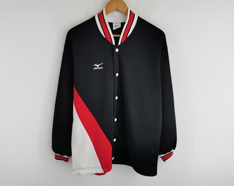 Puede incluir: Una chaqueta negra de manga larga con una franja diagonal blanca y roja en la parte delantera. La chaqueta presenta un logotipo de Mizuno blanco en el pecho, cierre frontal con botones y cuello y puños acanalados con rayas rojas y blancas.