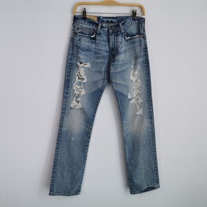 May include: Light blue denim jeans with distressed detailing, including rips and fading. The jeans are hanging on a wooden hanger against a white background. The jeans have a classic five-pocket design.