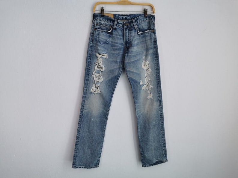 May include: Light blue denim jeans with distressed detailing, including rips and fading. The jeans are hanging on a wooden hanger against a white background. The jeans have a classic five-pocket design.