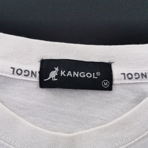 Peut inclure: Gros plan sur un col de t-shirt blanc avec une &eacute;tiquette noire portant le logo KANGOL et une silhouette de kangourou. L'&eacute;tiquette indique &eacute;galement la taille "M". La marque "KANGOL" est imprim&eacute;e sur le col.