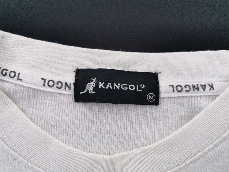 Peut inclure: Gros plan sur un col de t-shirt blanc avec une &eacute;tiquette noire portant le logo KANGOL et une silhouette de kangourou. L'&eacute;tiquette indique &eacute;galement la taille "M". La marque "KANGOL" est imprim&eacute;e sur le col.