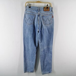 May include: Light blue denim jeans with a classic five-pocket design, featuring a straight leg and a faded wash. The jeans have a leather patch and a red logo tag. They are hanging on a gold-colored hanger against a white background.