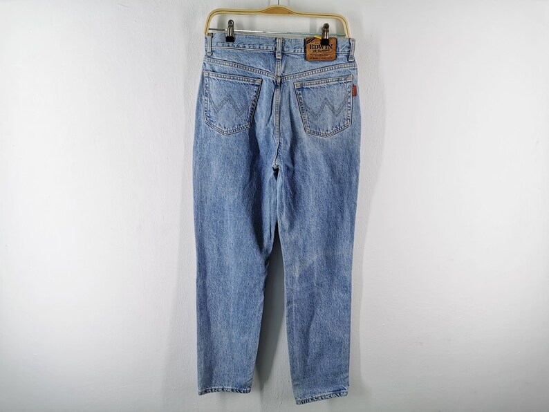 May include: Light blue denim jeans with a classic five-pocket design, featuring a straight leg and a faded wash. The jeans have a leather patch and a red logo tag. They are hanging on a gold-colored hanger against a white background.