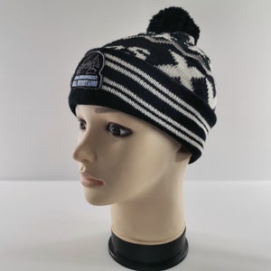 May include: A black and white knit beanie with a camouflage pattern and a black pom-pom. The hat features a patch with text. The brim has white stripes. The hat is displayed on a mannequin head.