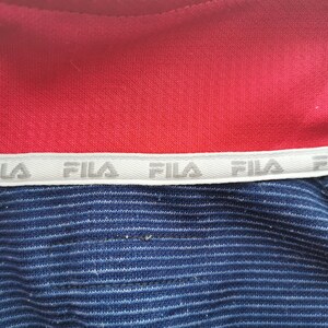 Puede incluir: Una prenda roja y azul con una banda interior blanca en el cuello que muestra el texto FILA repetido en color gris. La secci&oacute;n roja es de tejido liso, mientras que la secci&oacute;n azul presenta una textura finamente acanalada en horizontal.