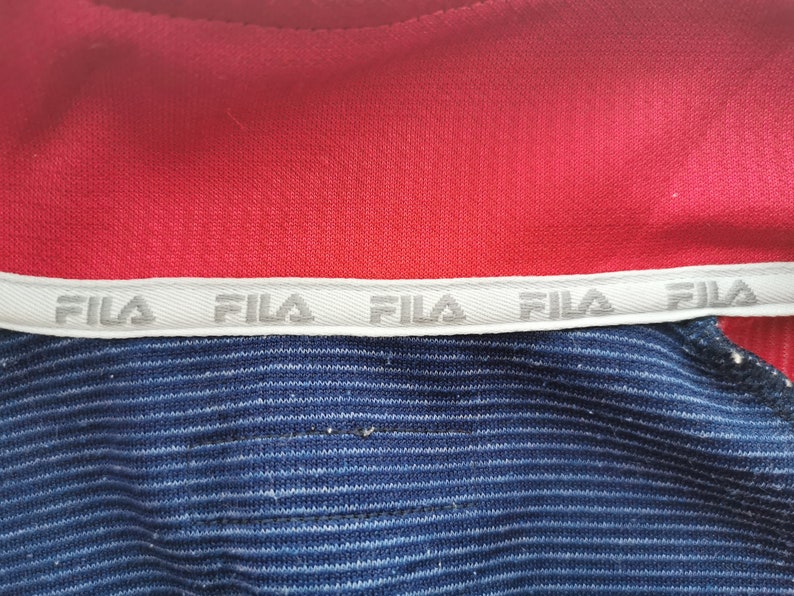 Puede incluir: Una prenda roja y azul con una banda interior blanca en el cuello que muestra el texto FILA repetido en color gris. La secci&oacute;n roja es de tejido liso, mientras que la secci&oacute;n azul presenta una textura finamente acanalada en horizontal.
