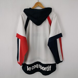 K&ouml;nnte beinhalten: Wei&szlig;-rot-marineblaue Kapuzenjacke mit dem Text "le coq sportif" am unteren Rand. Die Jacke hat eine marineblaue Kapuze und Akzente an den &Auml;rmeln. Die Jacke h&auml;ngt an einem Holzkleiderb&uuml;gel.