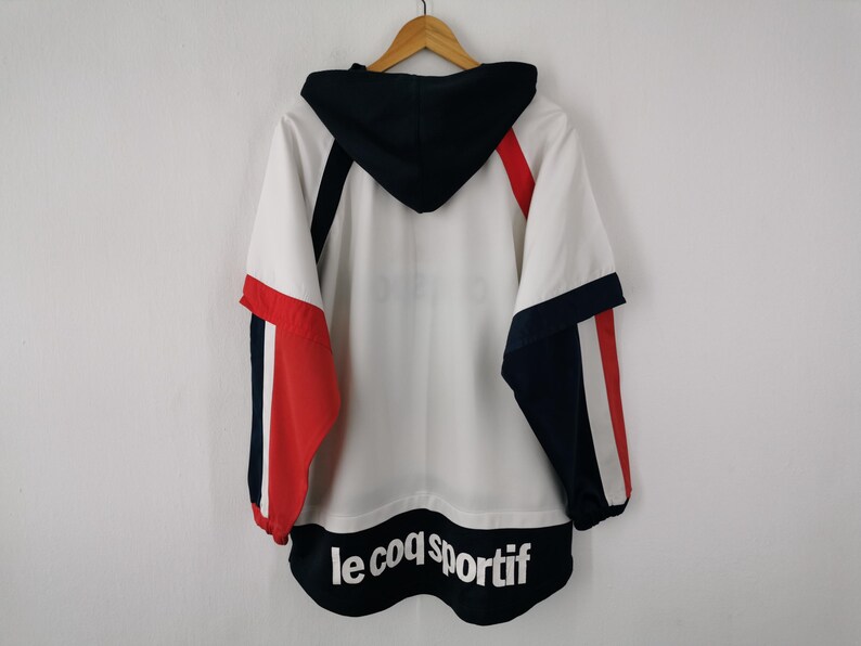 K&ouml;nnte beinhalten: Wei&szlig;-rot-marineblaue Kapuzenjacke mit dem Text "le coq sportif" am unteren Rand. Die Jacke hat eine marineblaue Kapuze und Akzente an den &Auml;rmeln. Die Jacke h&auml;ngt an einem Holzkleiderb&uuml;gel.