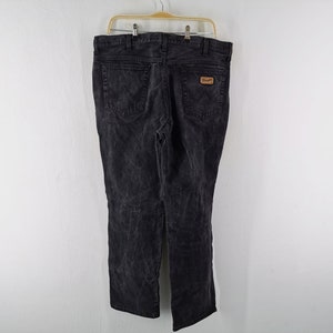 May include: Black denim jeans with a faded wash, hanging on a wooden hanger. The jeans have a classic five-pocket design and a brown leather patch with the Wrangler logo on the back pocket.