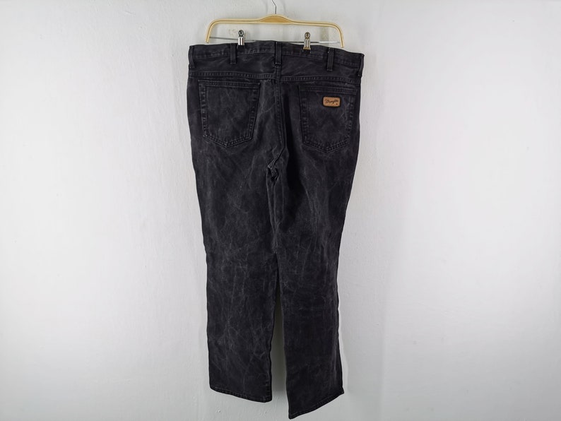 May include: Black denim jeans with a faded wash, hanging on a wooden hanger. The jeans have a classic five-pocket design and a brown leather patch with the Wrangler logo on the back pocket.
