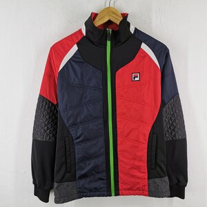 May include: A color-blocked track jacket in red, navy blue, black, and white. The jacket features a high collar, a full-length green zipper, and the Fila logo on the chest. The sleeves have a quilted pattern on the sides.
