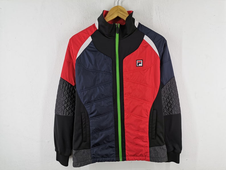 May include: A color-blocked track jacket in red, navy blue, black, and white. The jacket features a high collar, a full-length green zipper, and the Fila logo on the chest. The sleeves have a quilted pattern on the sides.