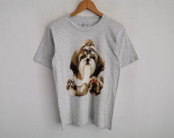 Puede incluir: Una camiseta gris jaspeado de manga corta con un estampado gráfico de tres perros Shih Tzu. El perro central tiene un lazo azul en el pelo, mientras que los perros más pequeños de abajo llevan lazos de color azul y rojo respectivamente.