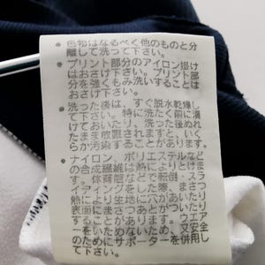 K&ouml;nnte beinhalten: Nahaufnahme eines wei&szlig;en Etiketts mit japanischem Text, das an einem marineblauen und wei&szlig;en Kleidungsst&uuml;ck befestigt ist. Das Etikett enth&auml;lt Pflegehinweise, einschlie&szlig;lich Wasch- und B&uuml;gelanweisungen.