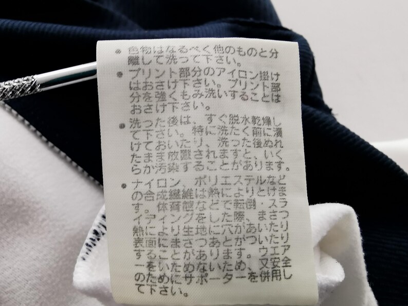 K&ouml;nnte beinhalten: Nahaufnahme eines wei&szlig;en Etiketts mit japanischem Text, das an einem marineblauen und wei&szlig;en Kleidungsst&uuml;ck befestigt ist. Das Etikett enth&auml;lt Pflegehinweise, einschlie&szlig;lich Wasch- und B&uuml;gelanweisungen.