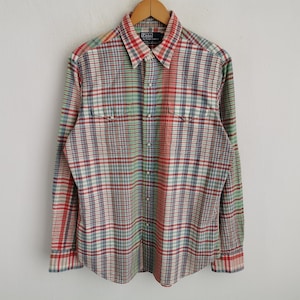 May include: A long-sleeved plaid shirt in red, green, blue, and white. The shirt has a button-down front, two chest pockets, and a collar. The shirt is hanging on a wooden hanger.