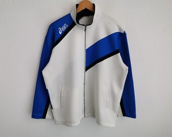 Puede incluir: Una chaqueta deportiva con cremallera en blanco, azul y negro, con cuello alto y mangas largas. El logotipo de Asics aparece en blanco en la parte superior derecha del pecho. El diseño incluye paneles diagonales de colores.