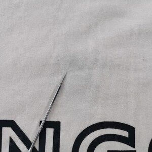 Peut inclure: T-shirt cr&egrave;me avec un motif graphique noir. Une aiguille argent&eacute;e est au premier plan, pointant vers le t-shirt. Le tissu du t-shirt a un aspect l&eacute;g&egrave;rement textur&eacute;.