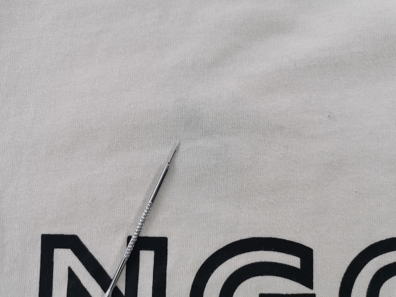 Peut inclure: T-shirt cr&egrave;me avec un motif graphique noir. Une aiguille argent&eacute;e est au premier plan, pointant vers le t-shirt. Le tissu du t-shirt a un aspect l&eacute;g&egrave;rement textur&eacute;.