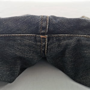 May include: Close-up of dark wash denim jeans. The image focuses on the waistband and the seam details, showcasing the stitching in a contrasting thread colour. The fabric has a textured appearance.