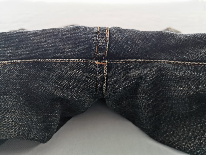May include: Close-up of dark wash denim jeans. The image focuses on the waistband and the seam details, showcasing the stitching in a contrasting thread colour. The fabric has a textured appearance.