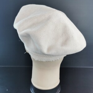 Puede incluir: Un boina de color beige claro hecho de tejido de punto. El sombrero se exhibe en una cabeza de maniqu&iacute;. El sombrero tiene una corona redondeada y un ala peque&ntilde;a. El tejido tiene una textura sutil.