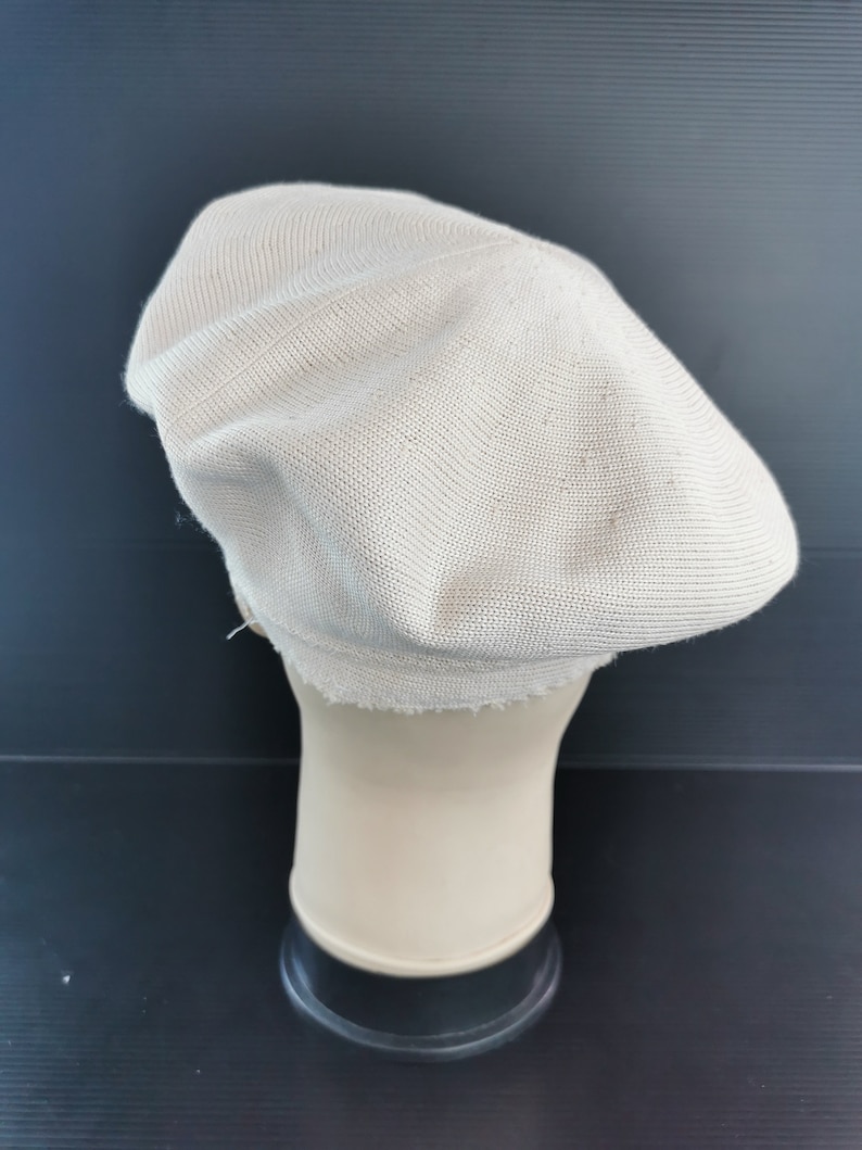 Puede incluir: Un boina de color beige claro hecho de tejido de punto. El sombrero se exhibe en una cabeza de maniqu&iacute;. El sombrero tiene una corona redondeada y un ala peque&ntilde;a. El tejido tiene una textura sutil.