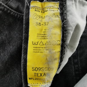 May include: Close-up of a yellow Wrangler jeans tag with black text and a black denim background. The tag reads "Wrangler 38-32" and includes washing instructions. The jeans are a dark gray wash.