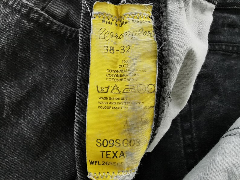 May include: Close-up of a yellow Wrangler jeans tag with black text and a black denim background. The tag reads "Wrangler 38-32" and includes washing instructions. The jeans are a dark gray wash.