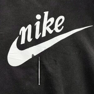 K&ouml;nnte beinhalten: Nahaufnahme eines schwarzen Kleidungsst&uuml;cks mit dem wei&szlig;en Nike-Logo und Swoosh. Das Wort "nike" ist in einer fetten, stilisierten Schriftart gehalten. Unter dem Logo befindet sich ein silbernes Werkzeug.