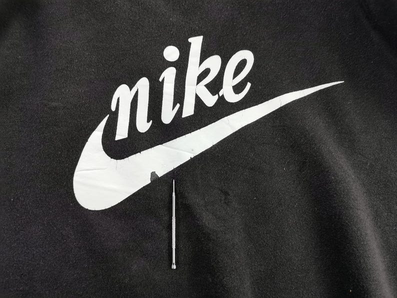 K&ouml;nnte beinhalten: Nahaufnahme eines schwarzen Kleidungsst&uuml;cks mit dem wei&szlig;en Nike-Logo und Swoosh. Das Wort "nike" ist in einer fetten, stilisierten Schriftart gehalten. Unter dem Logo befindet sich ein silbernes Werkzeug.