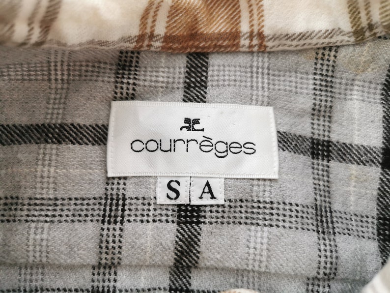 May include: Close-up of a plaid shirt with a label that reads "courr&egrave;ges" in black text. The shirt has a gray, white, and brown plaid pattern. The label also has the size "S A" printed on it.