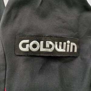 Pode incluir: Casaco preto com o patch prateado do log&oacute;tipo "GOLDWIN". O patch &eacute; retangular e cosido no casaco. O casaco &eacute; preto com uma risca vermelha vis&iacute;vel na lateral.