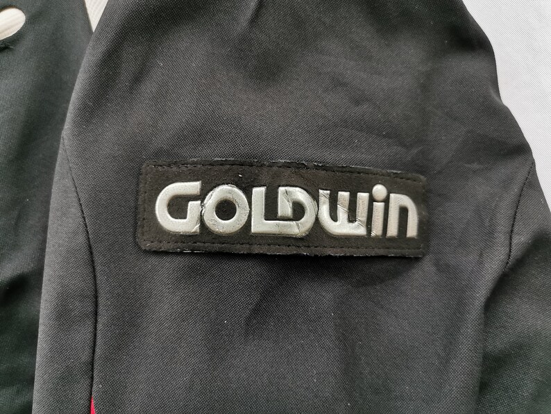 Pode incluir: Casaco preto com o patch prateado do log&oacute;tipo "GOLDWIN". O patch &eacute; retangular e cosido no casaco. O casaco &eacute; preto com uma risca vermelha vis&iacute;vel na lateral.