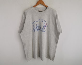 Puede incluir: Una camiseta gris jaspeado de manga corta con un gráfico de Eeyore tumbado con un lazo rosa en la cola. El texto de arriba dice "Happiness is in the little things." y el nombre "Eeyore" está impreso en azul debajo del personaje.