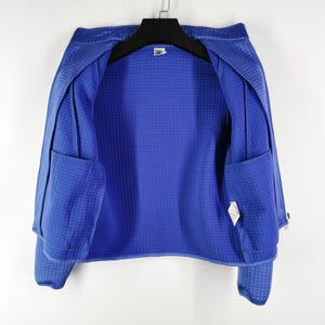 May include: A vibrant blue jacket with a textured, grid-like pattern. The jacket features two front pockets and is hanging on a black hanger. The interior lining is a solid blue, and the jacket appears to be made of a lightweight material.