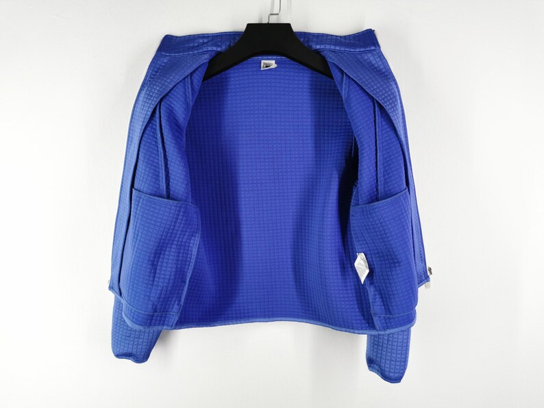 May include: A vibrant blue jacket with a textured, grid-like pattern. The jacket features two front pockets and is hanging on a black hanger. The interior lining is a solid blue, and the jacket appears to be made of a lightweight material.