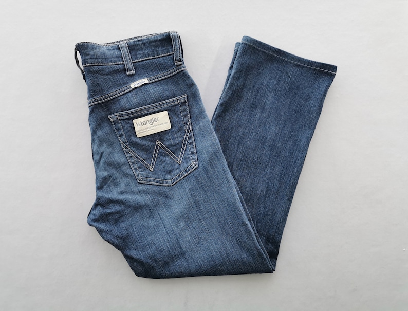 May include: Blue denim Wrangler jeans with a classic five-pocket design. The jeans have a straight leg, a button and zip closure, and the Wrangler logo on the back pocket and waistband. The jeans are neatly folded.
