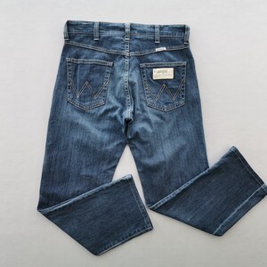 May include: Blue denim jeans with the Wrangler logo on the back pockets and waistband. The jeans have a classic five-pocket design and a straight leg fit. The jeans are made of a durable denim fabric.