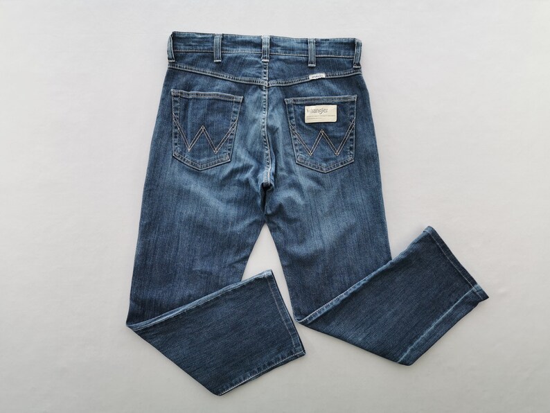 May include: Blue denim jeans with the Wrangler logo on the back pockets and waistband. The jeans have a classic five-pocket design and a straight leg fit. The jeans are made of a durable denim fabric.