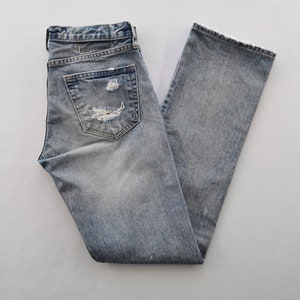 May include: Light blue denim jeans with distressed details, including a ripped pocket and a frayed hem. The jeans are folded and laid flat, showcasing their faded wash and classic five-pocket design.